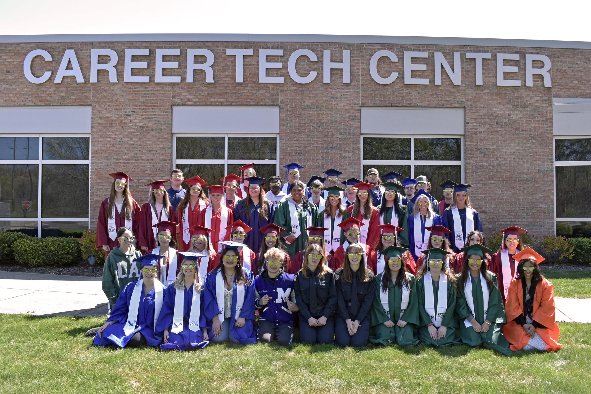 A large group of graduating students in colorful caps and gowns pose together outside the Career Tech Center building.