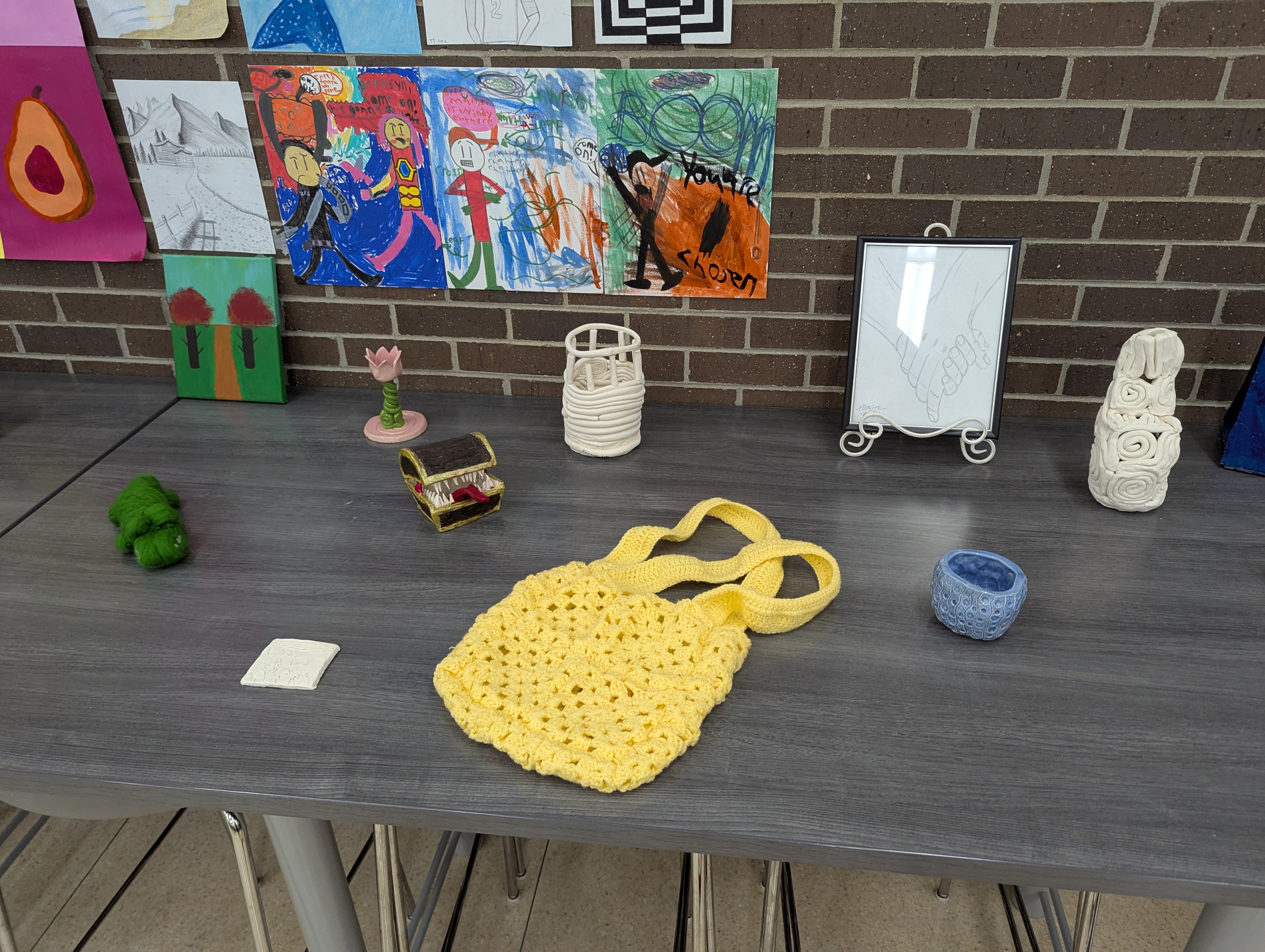 A student art display with paintings and drawings hung on a brick wall above a table holding ceramic pieces, a framed sketch, and a yellow crocheted bag.