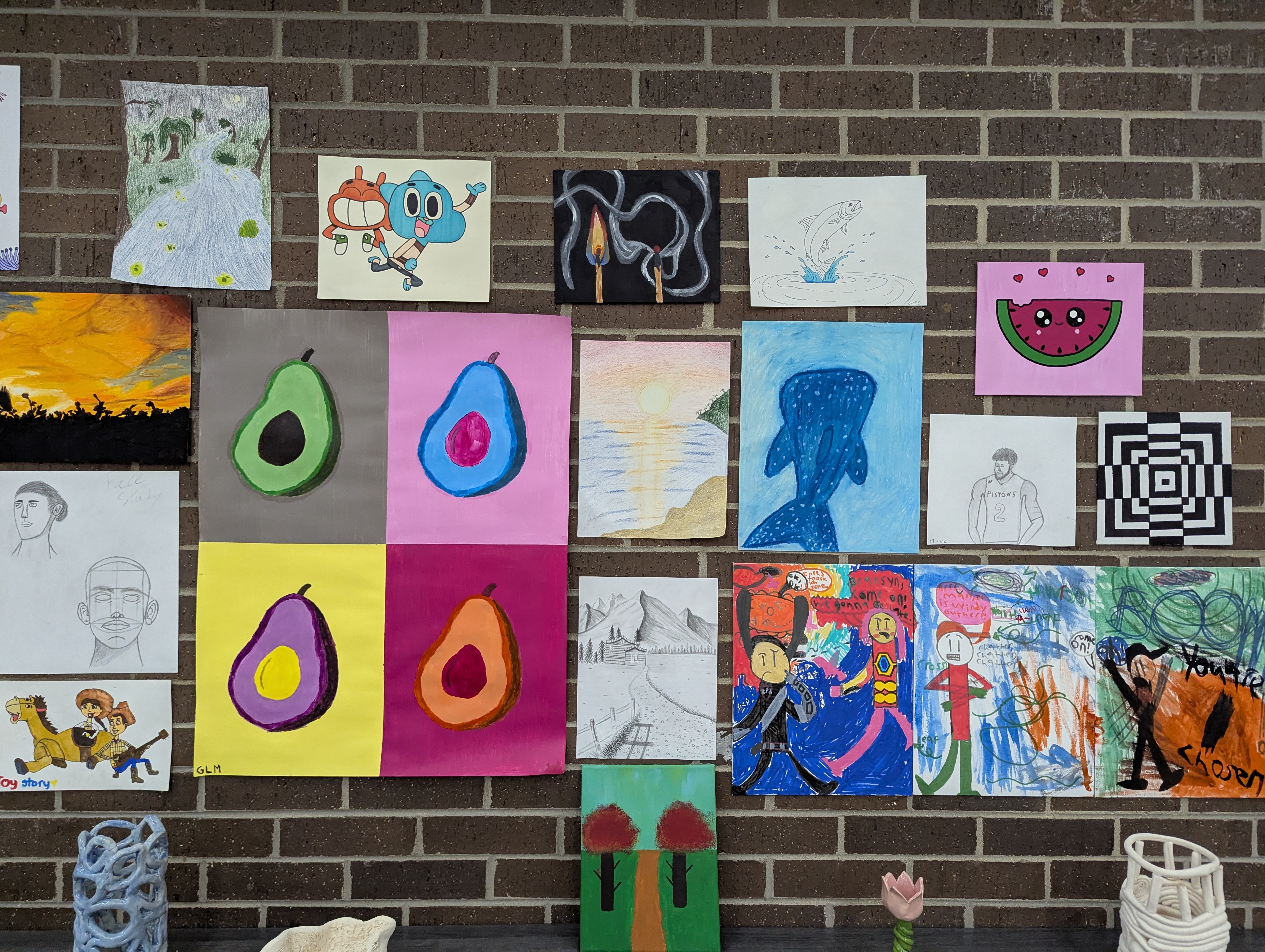 A student art display on a brick wall with paintings, drawings, and pop art style avocados, plus ceramic pieces on a shelf below.