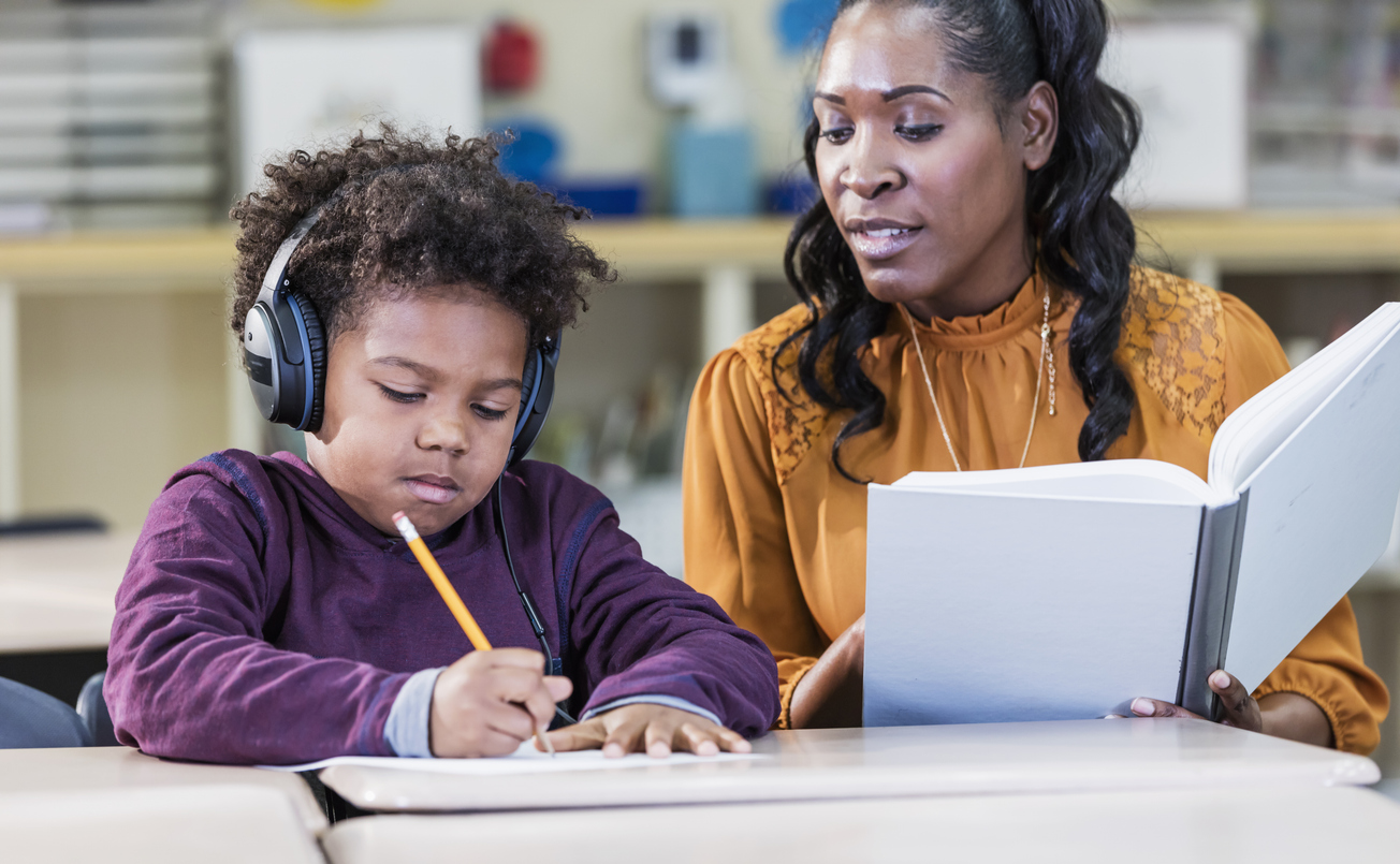 An African-American teacher with an elementary school student in the classroom. The student is an 8 year old boy. Noise canceling headphones can be helpful to a child with learning difficulty.