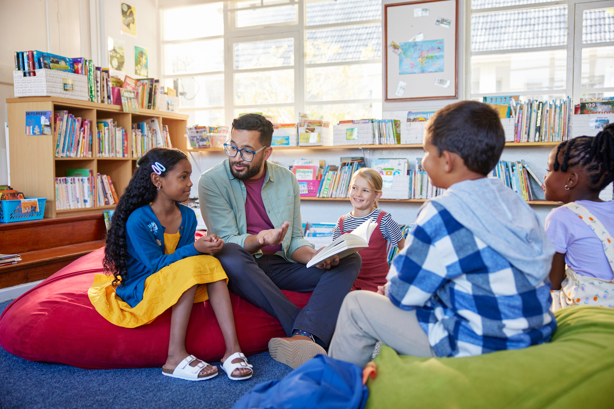 Male teacher reading a book to students while sitting in school library. Librarian read a fairy tale to a group of multiethnic primary schoolchildren. Middle eastern man at elementary school reading stories to little boys and curious girls.