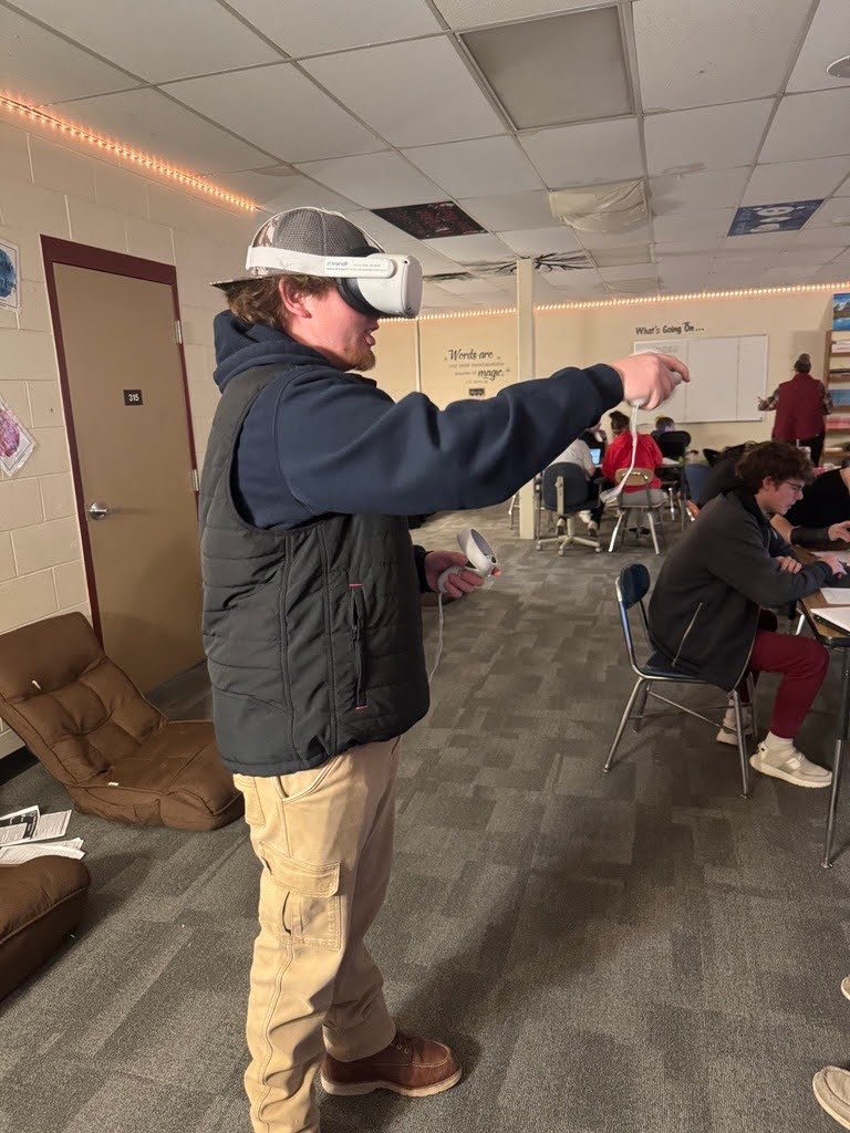 A student in a black vest, tan pants, and a baseball cap uses a VR headset and controllers in a classroom. He is standing near brown floor chairs, pointing with one hand while holding a controller in the other. Other students are seated at tables in the background, working or observing.