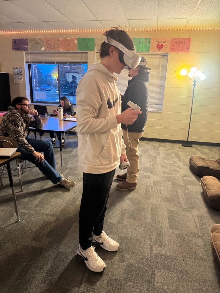A student wearing a white hoodie and black athletic pants uses a VR headset and handheld controller in a classroom setting. Behind him, other students are seated at desks working on laptops or talking. The room is warmly lit with string lights and posters on the wall that say “Empathetic” and “Respect.”