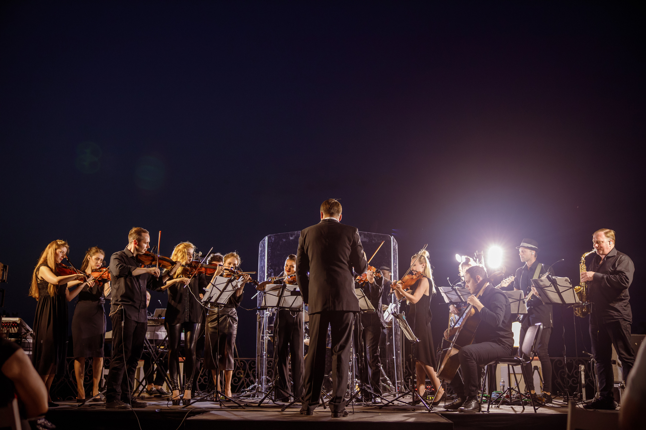 A conductor leads an orchestra of violinists, a cellist, and a saxophonist performing on an outdoor stage at night.