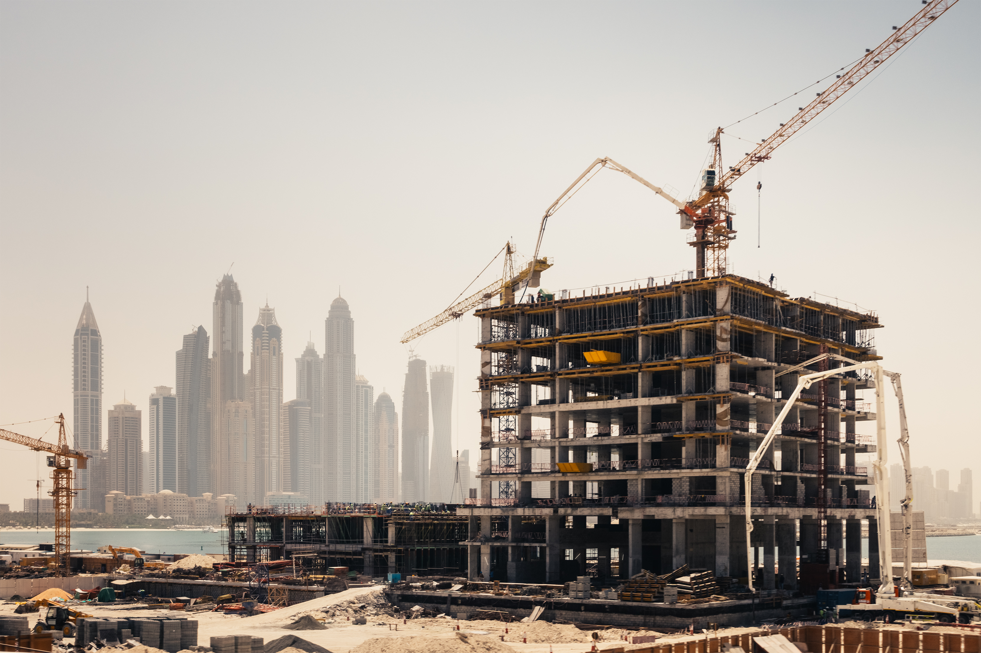 A concrete high rise under construction with cranes and concrete pumps, set against a hazy skyline of coastal skyscrapers.